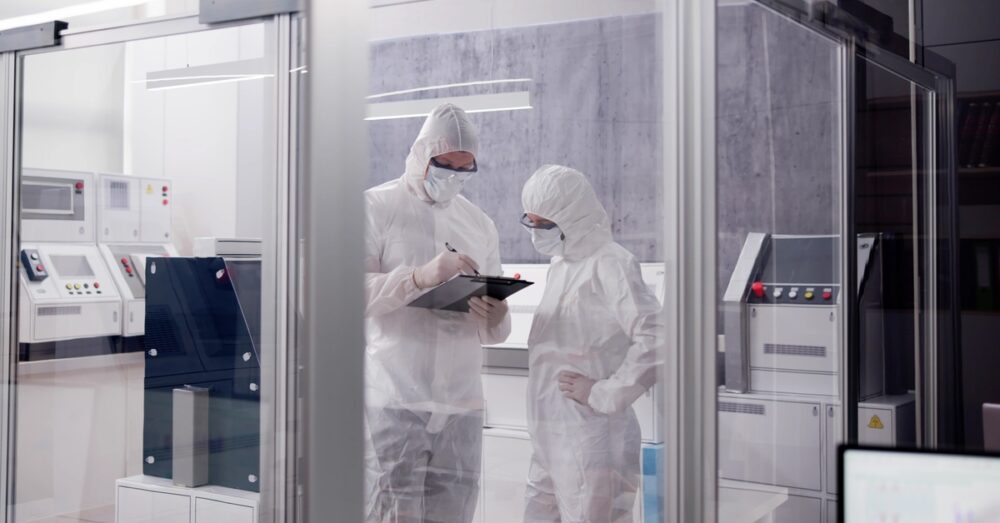 Two people stand in a laboratory space in all-white, full-body protective suits behind glass walls, inspecting a clipboard.